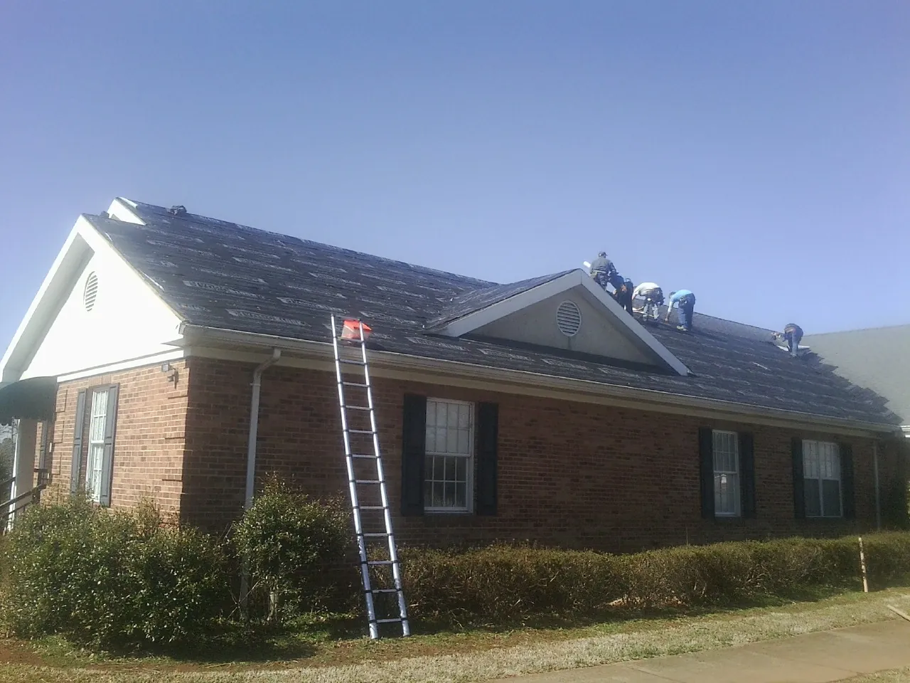 Crew tearing shingles off a brick ranch roof with a ladder set — Emergency Roof Repair work by our crew in Beech Grove