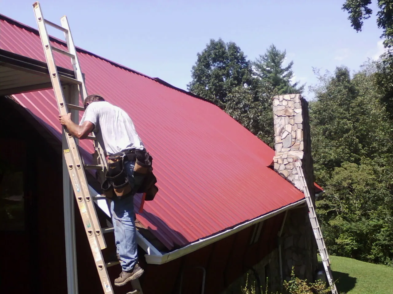 Roofer on a ladder beside a red metal roof and stone chimney in Beech Grove